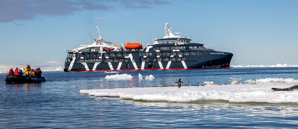 Le Magellan Explorer devant un iceberg avec un manchot dessus et un zodiac qui l'observe