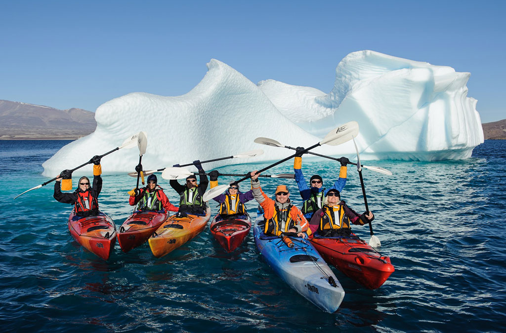 groupe de kayakiste qui levent leurs rames devant un iceberg pour la photo