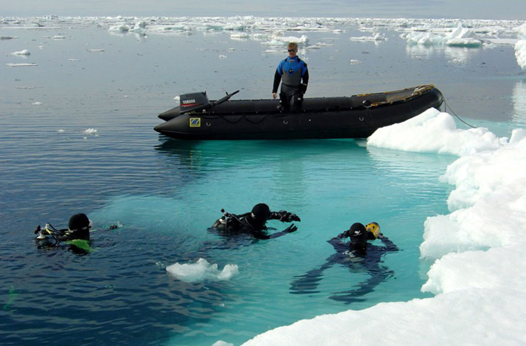 trois ppngeurs en antarctique à la surface de l'eau avec le zodiac qui surveille